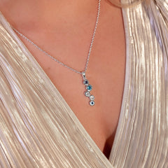 A close-up shot of a woman wearing the sterling silver topaz stream pendant, a matching bracelet, and a ring. She is wearing a shiny, pleated gold top and her hand is resting on her collarbone.