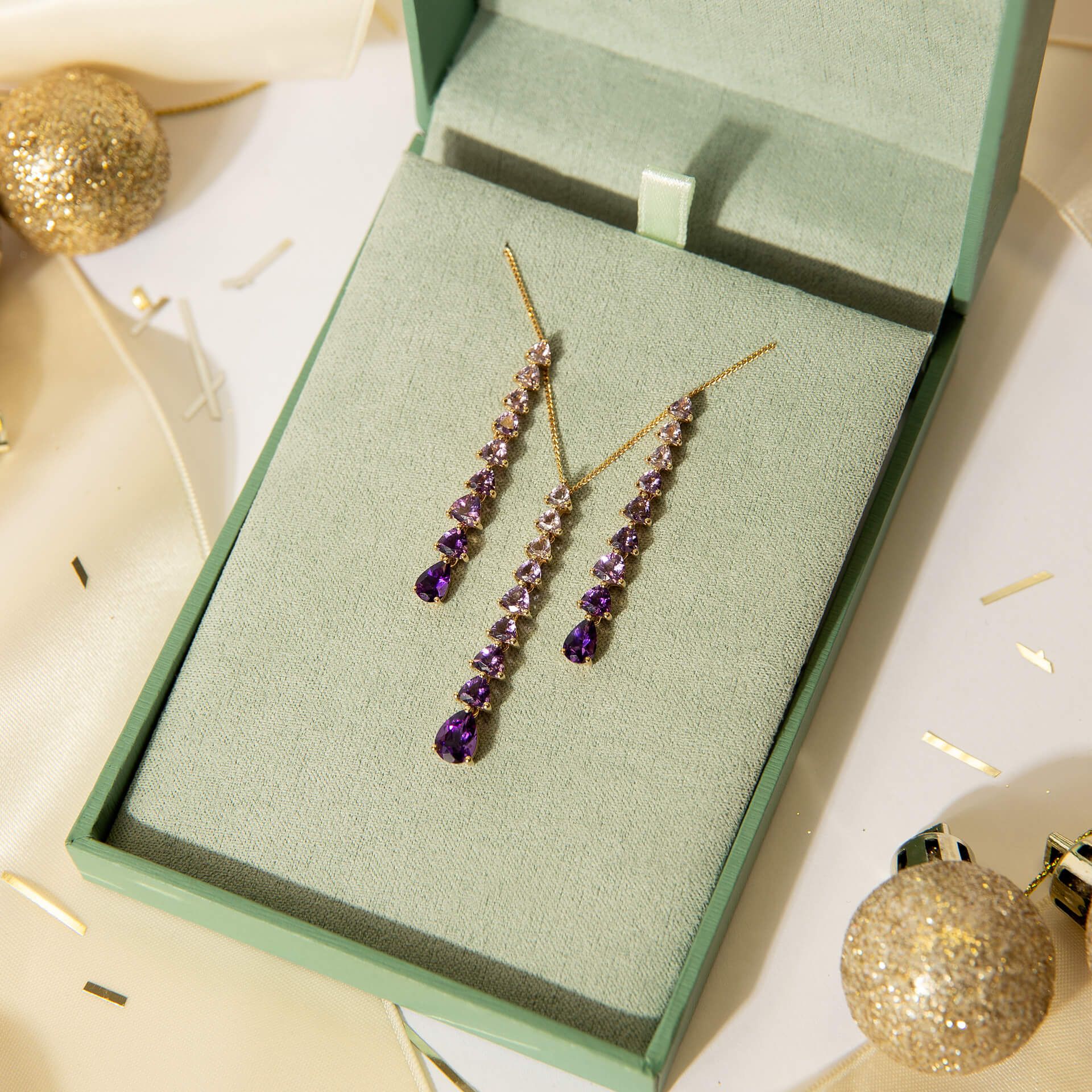 A 9ct gold amethyst pendant and earring set displayed in a green Elements Gold gift box, surrounded by gold Christmas baubles.