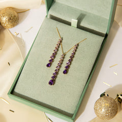A 9ct gold amethyst pendant and earring set displayed in a green Elements Gold gift box, surrounded by gold Christmas baubles.