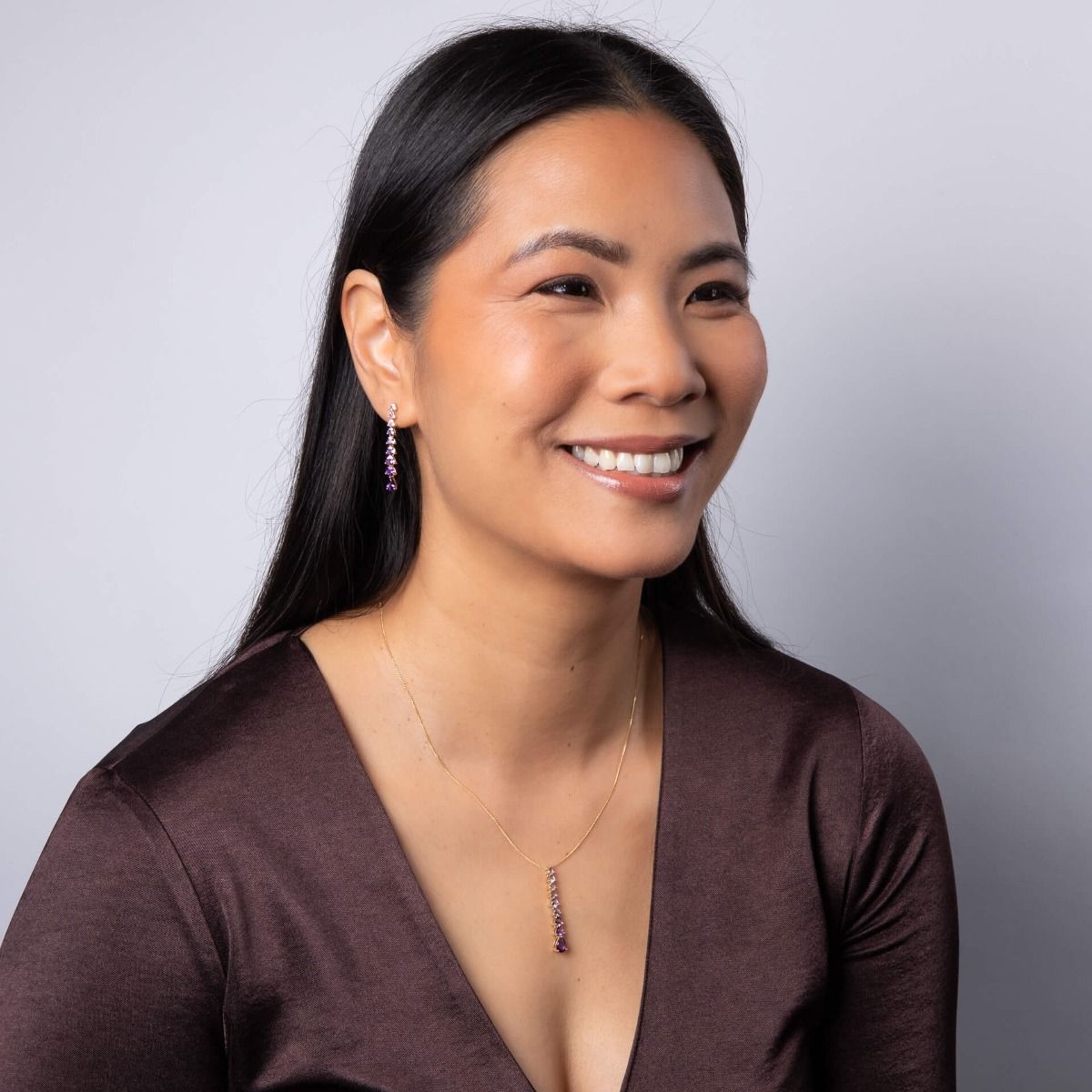 A smiling woman wearing a gold amethyst pendant necklace and matching drop earrings, dressed in a gold top with a Christmas tree in the background.